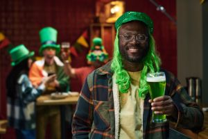 Happy young African American man in green cap and wig toasting with beer
