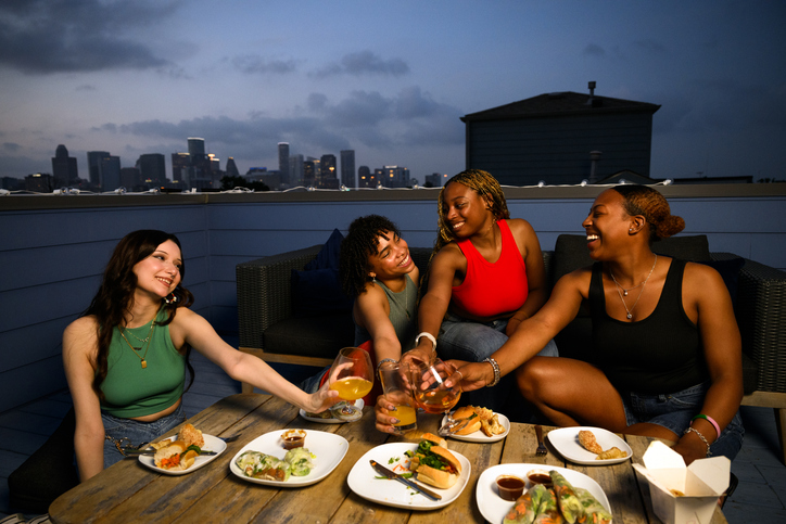 A group of girlfriends perform celebratory toast to friendship and togetherness over take out food on a blue rooftop with skyscrapers at dusk in background