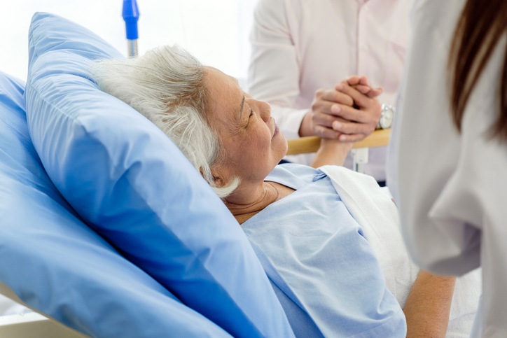 An elderly woman lies in a hospital bed, receiving comfort and support from a doctor and a family member. The scene captures healthcare, senior patient care, and emotional support in a medical setting