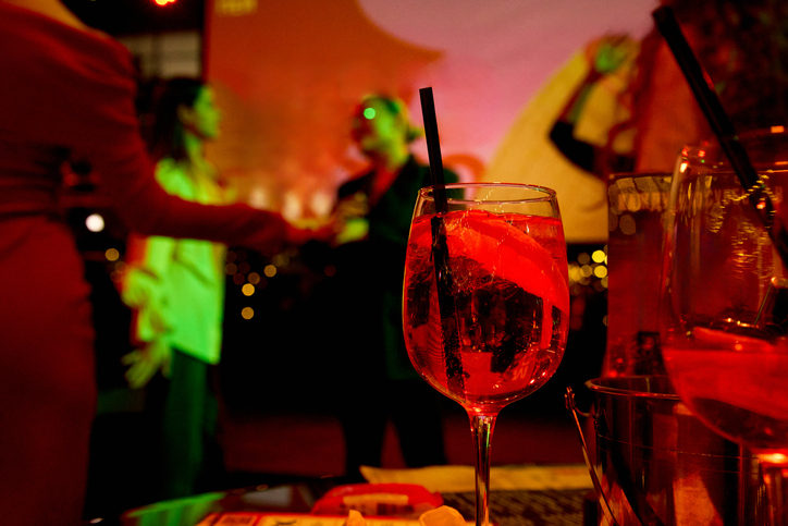 Close‑up of Aperol Spritz cocktail glass with dancing women in party background, festive design for Christmas, New Year, Birthday, Women’s Day, and alcohol‑free lifestyle concepts.